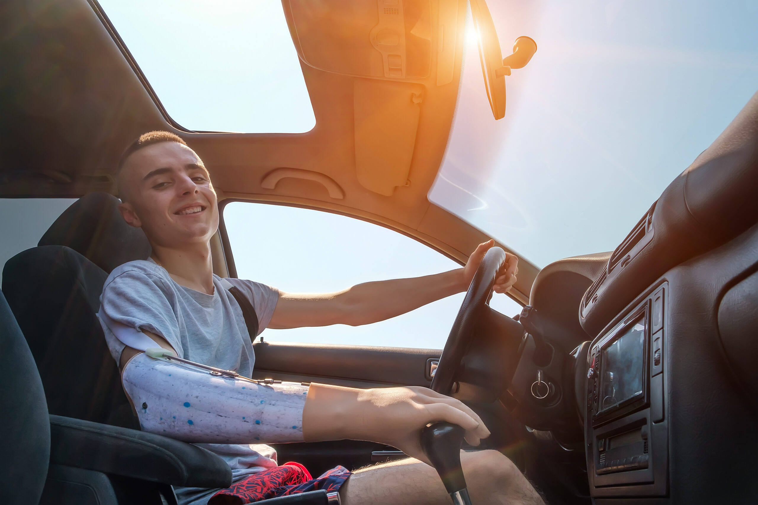 Young smiling caucasian man with amputated arm and prosthesis drives a car on a sunny day in summer. Side view inside of cabin