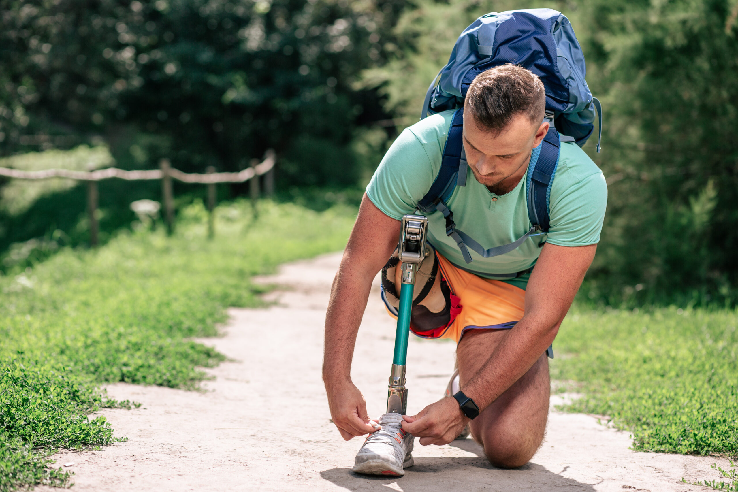 Person with a lower-limb prosthesis adjusting their shoe on an outdoor pathway.